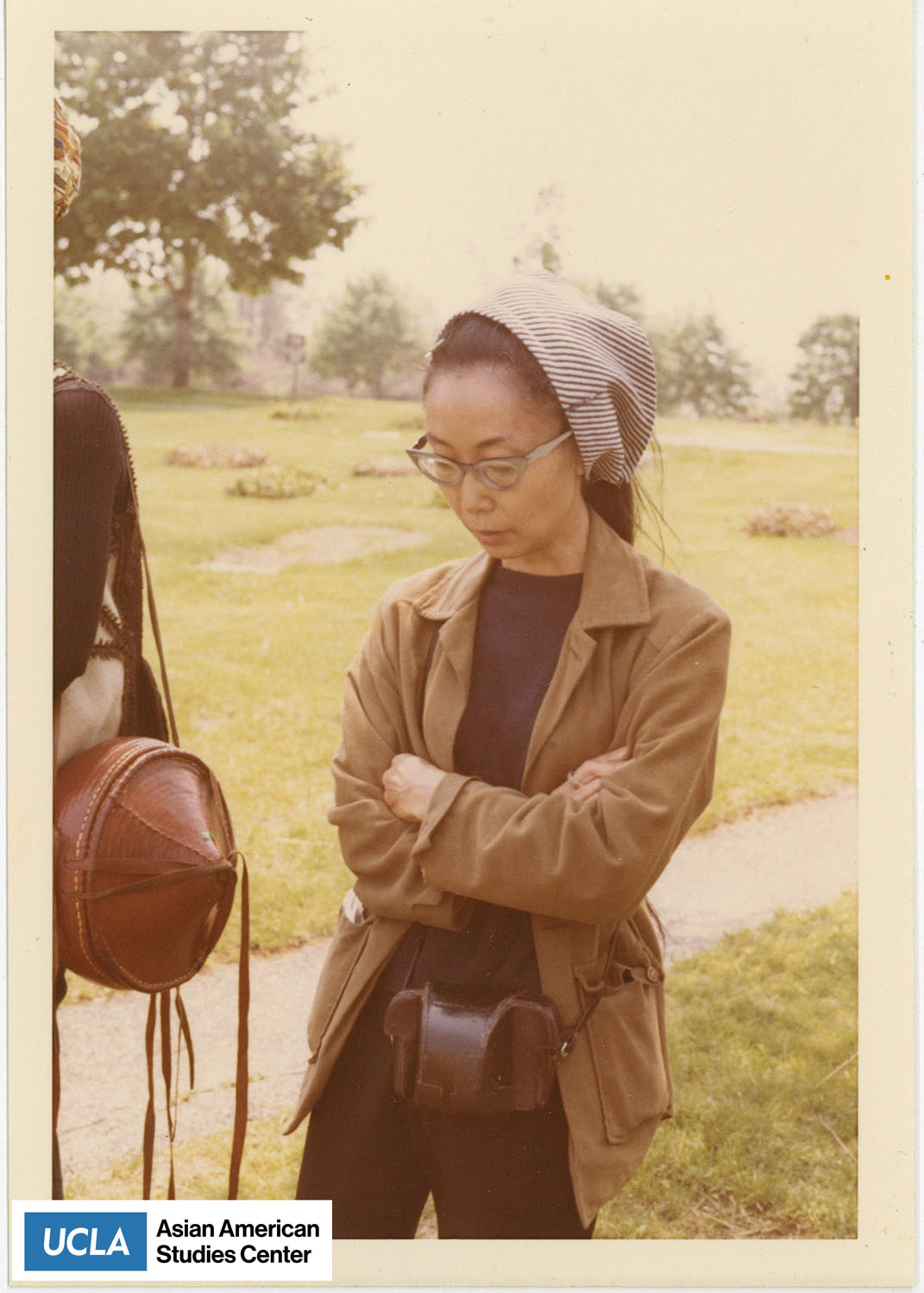 Photo of Yuri looking down with arms crossed. Yuri is wearing a brown jacket with a leather camera bag.