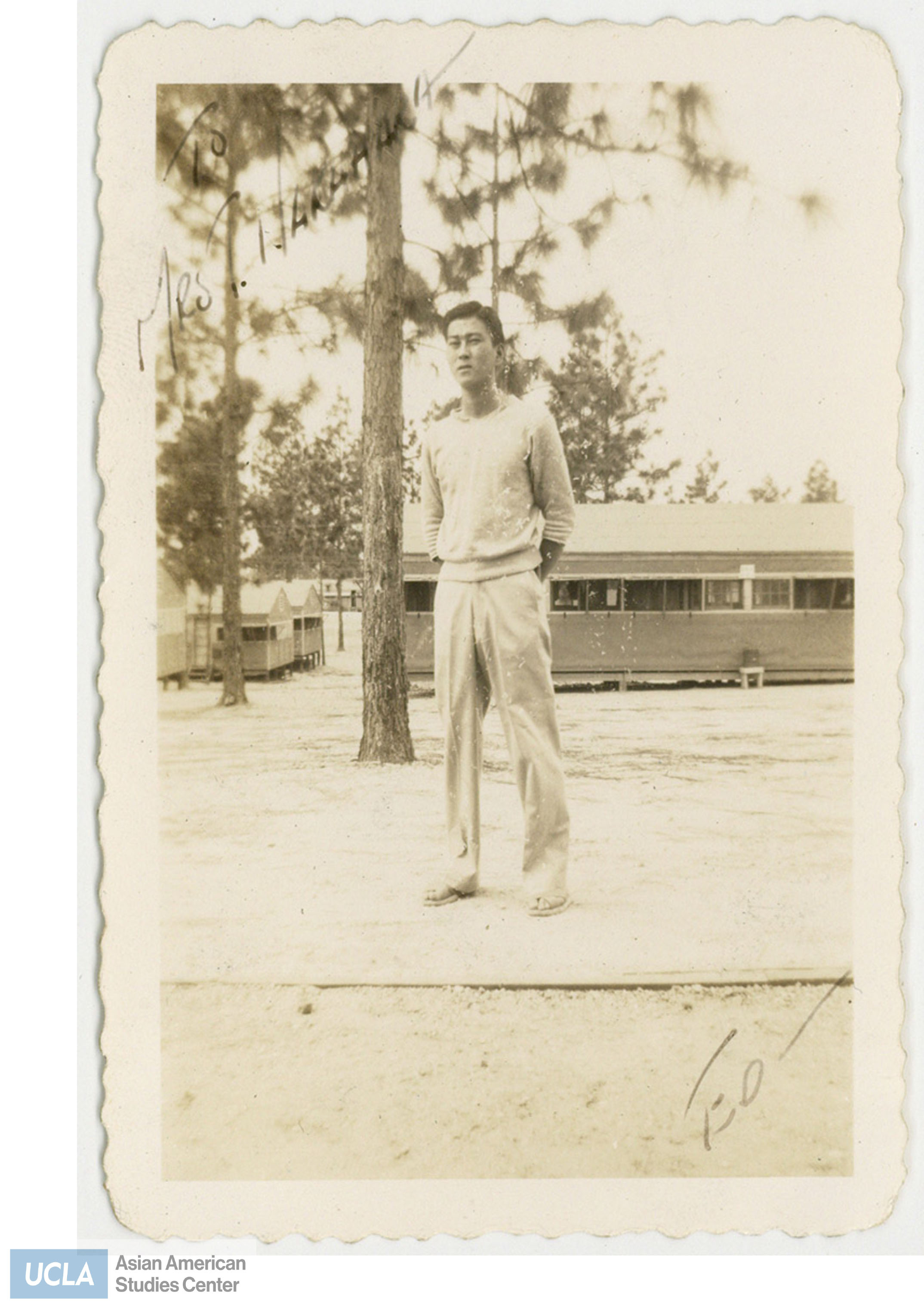 Ed standing infront of Jerome barracks and wearing a sweater and slacks. Back of photo includes: 