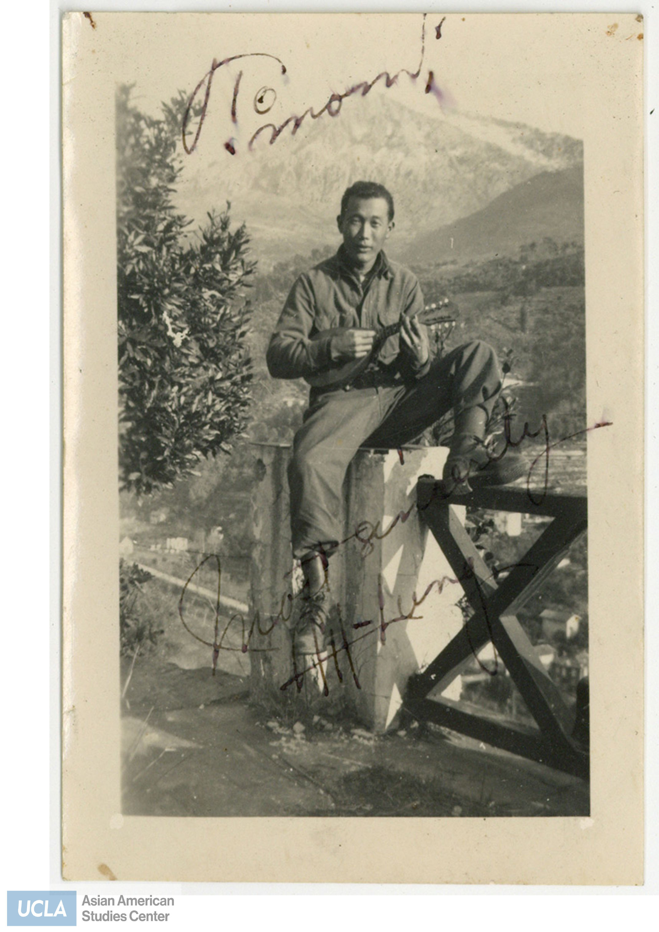 Nisei Solider sitting on fence and playing a mandolin. Back of photo includes text: 