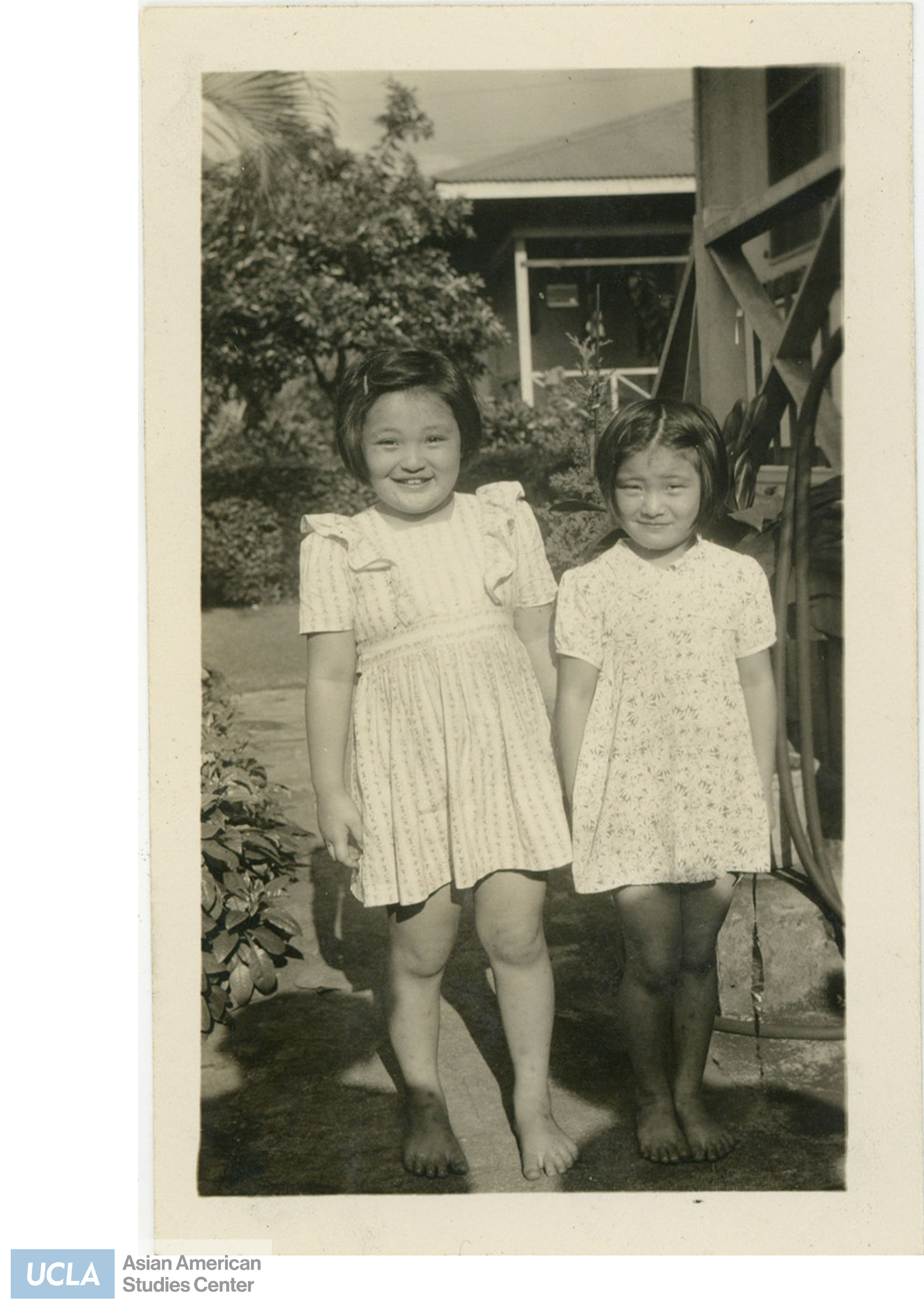 Two girls wearing dresses stand next to each other. Back of photo includes text: 