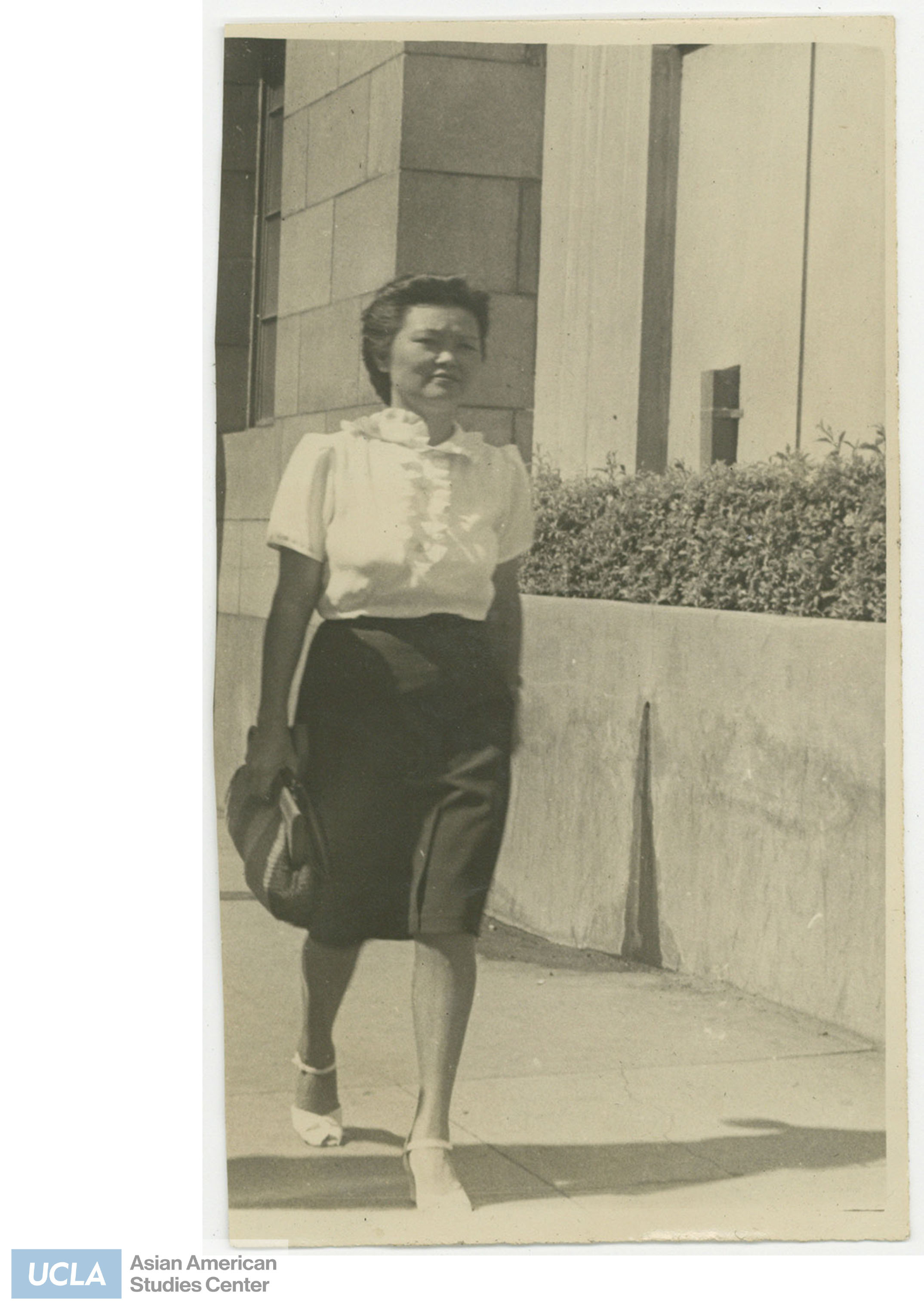 Photo of a woman wearing a blouse and skirt while walking on the sidewalk. Back of photo includes text: 