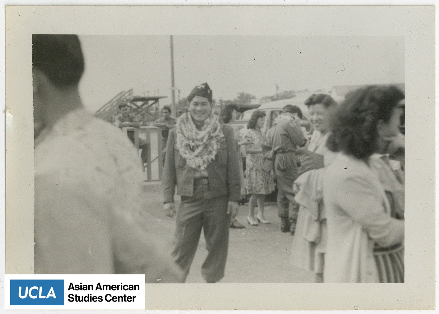 A smiling Nisei serviceman wearing multiple leis with people surrounding him.
