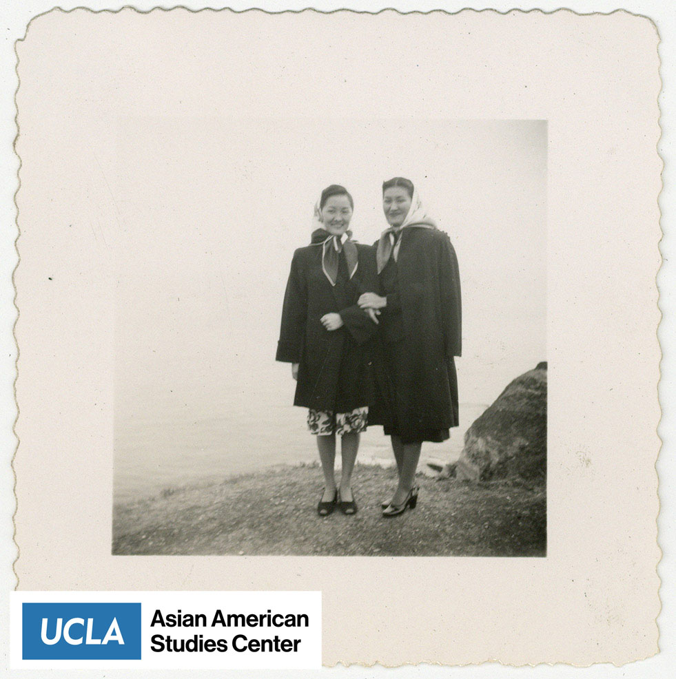 Two Nisei women with scarves wrapped around their heads stand infront of a body of water.