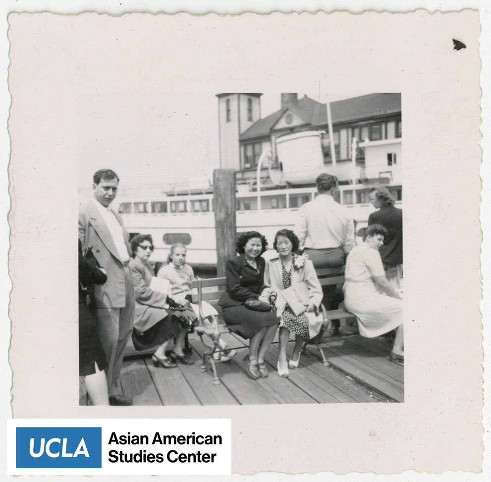 Tsuma and friend sit on a bench at the pier with a crowd of people.