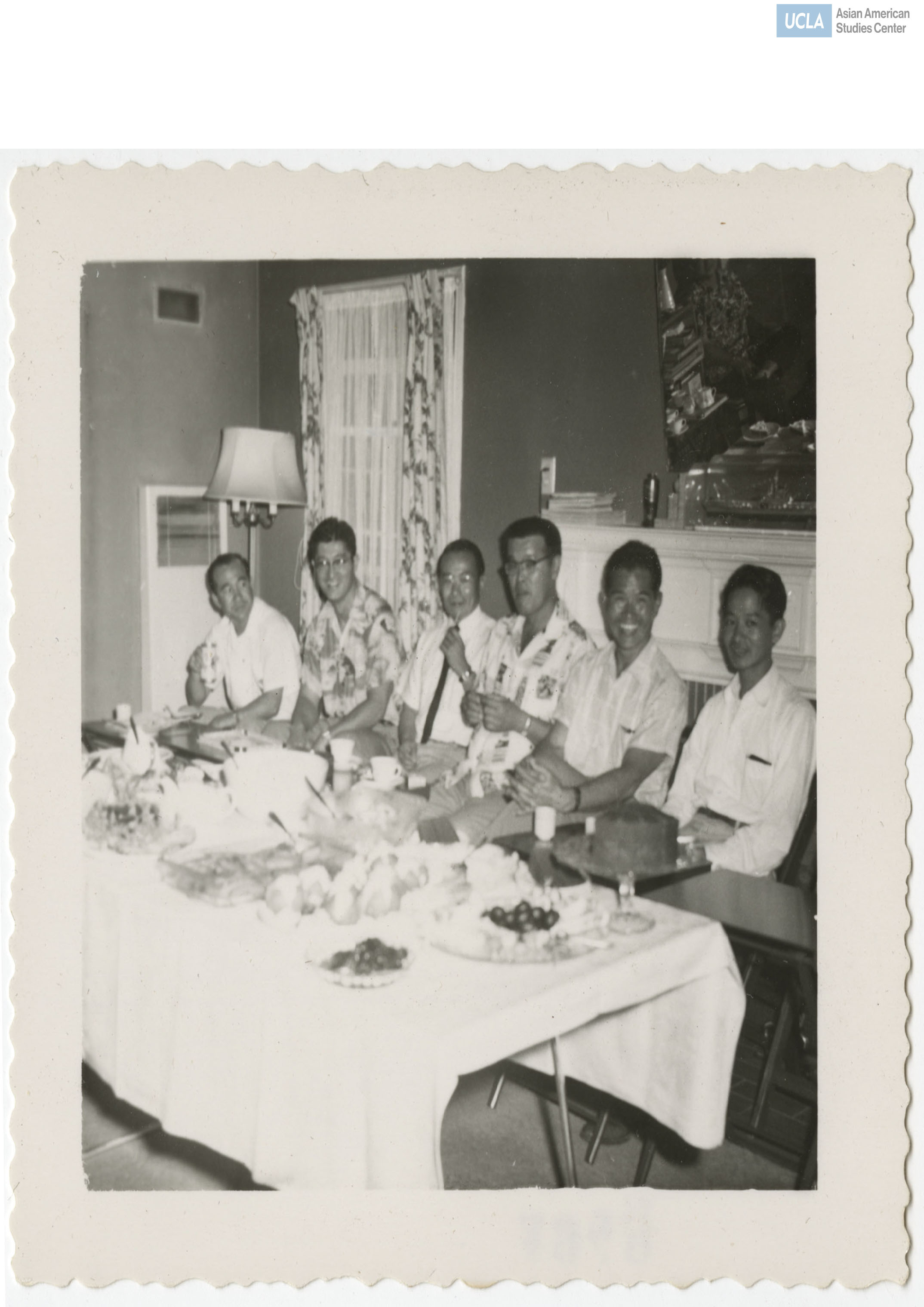 Photograph of six men sitting in a row during a celebration with food and drinks.
