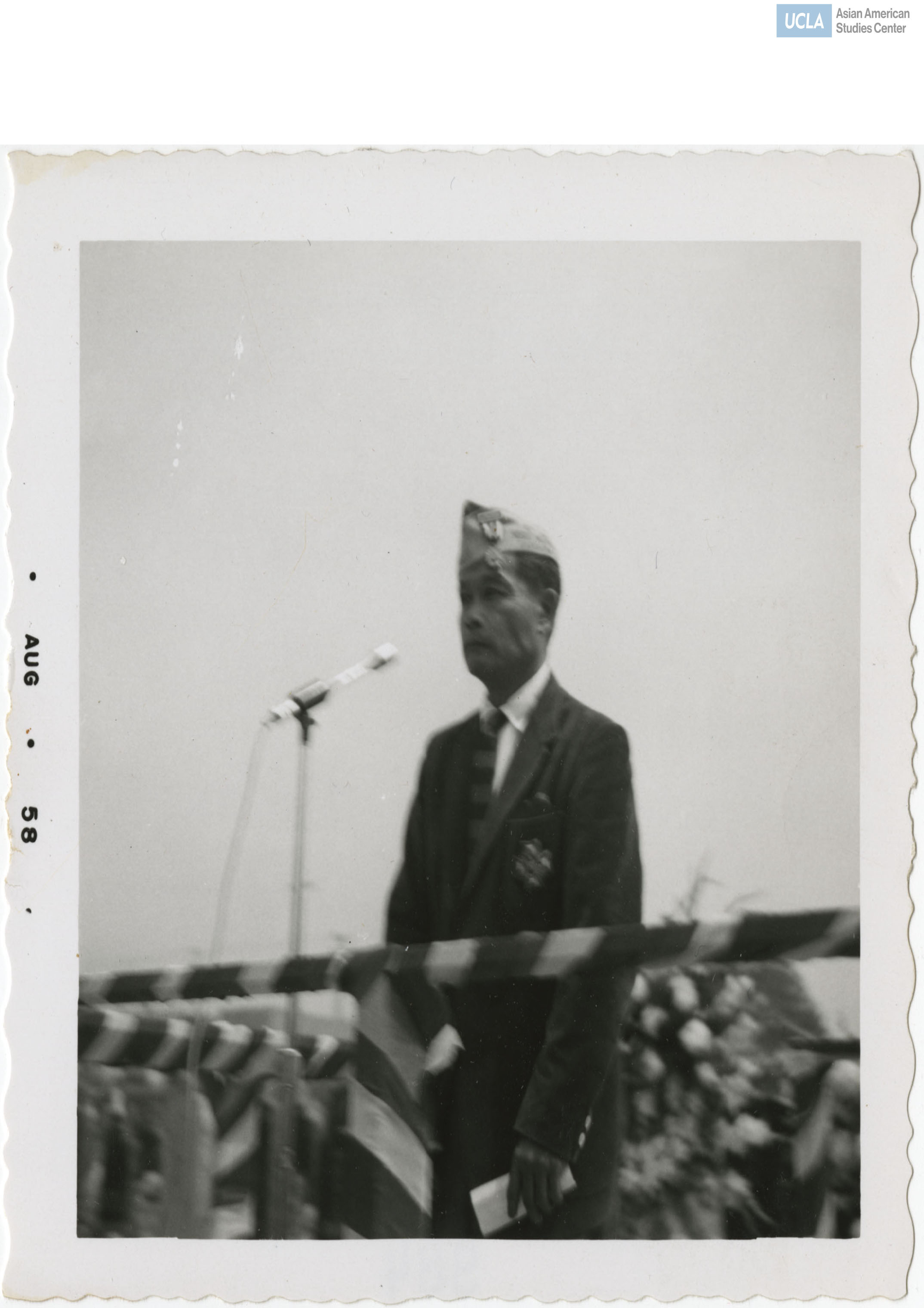 442nd Regimental Combat Team veteran in uniform wearing a garrison cap with a metal adornment stands before a microphone.