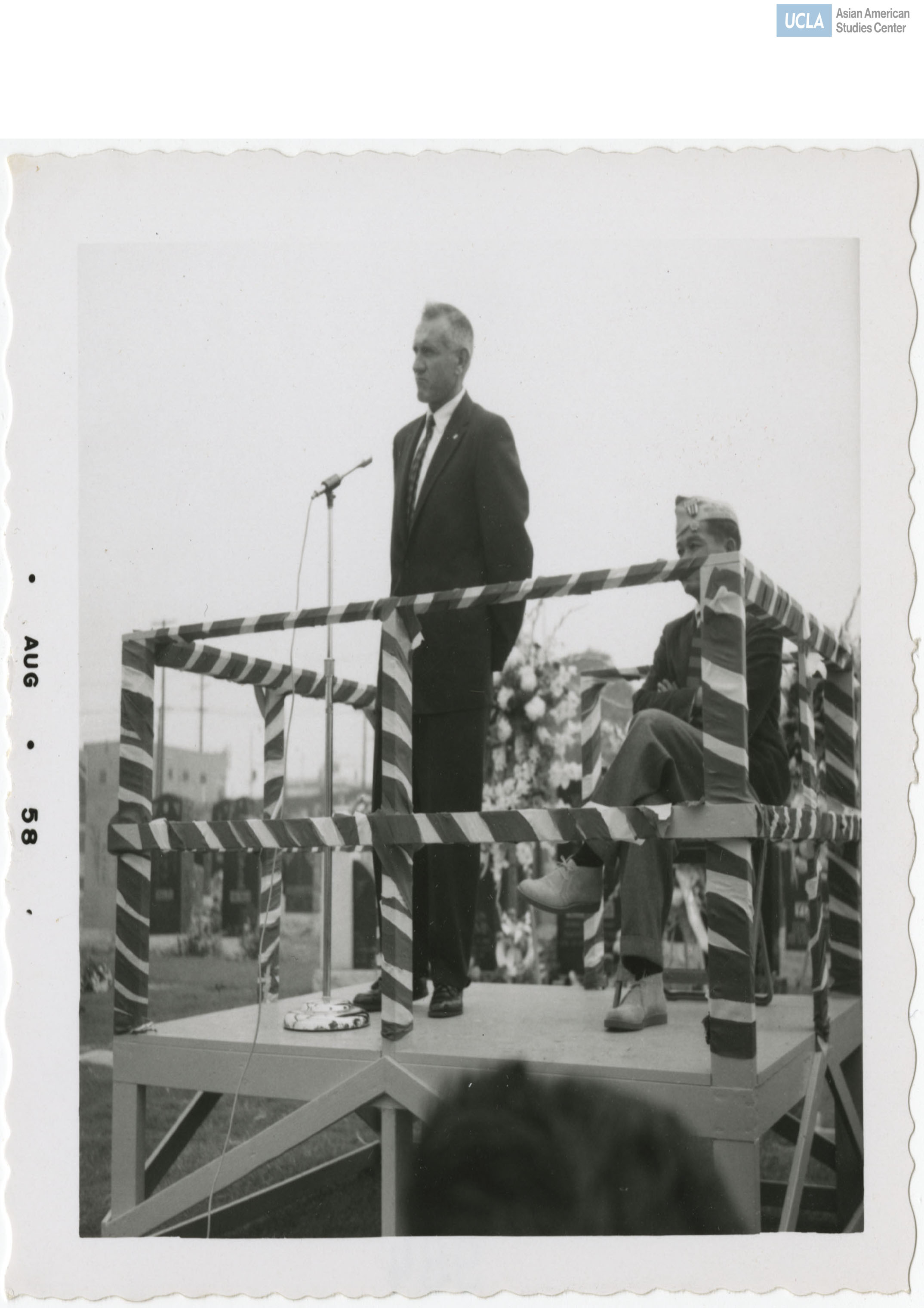 Man in suit with a decorated veteran on a raised stage during the 442nd Regimental Combat Team reunion.