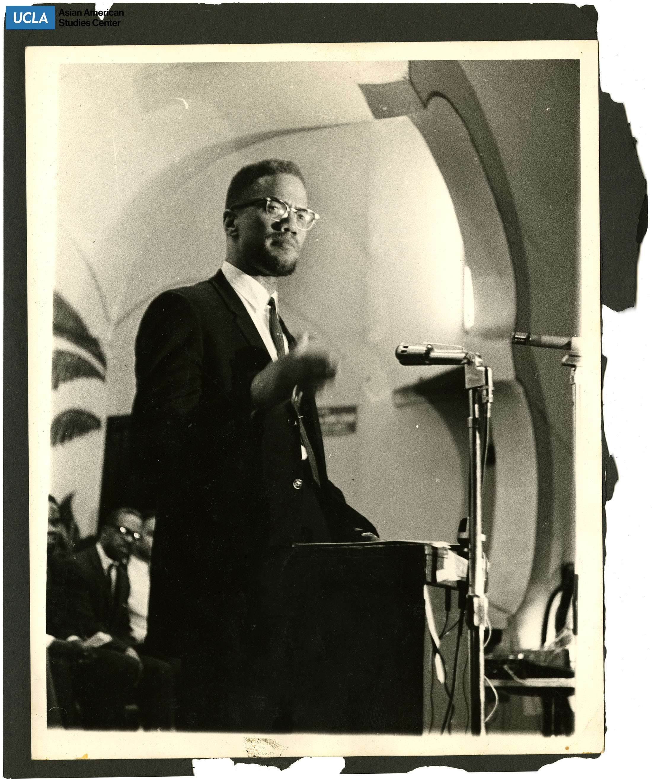 Malcolm X standing before a mircophone and podium.