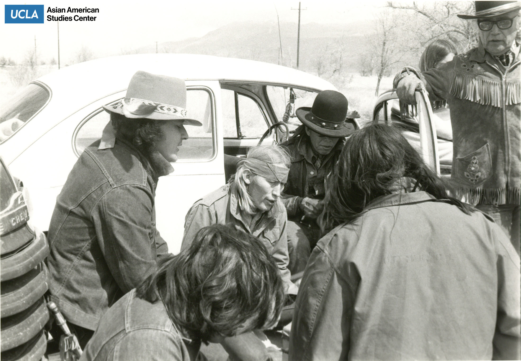 Black and white photograph of men sitting in a circle with Volkswagen Beetle in background.