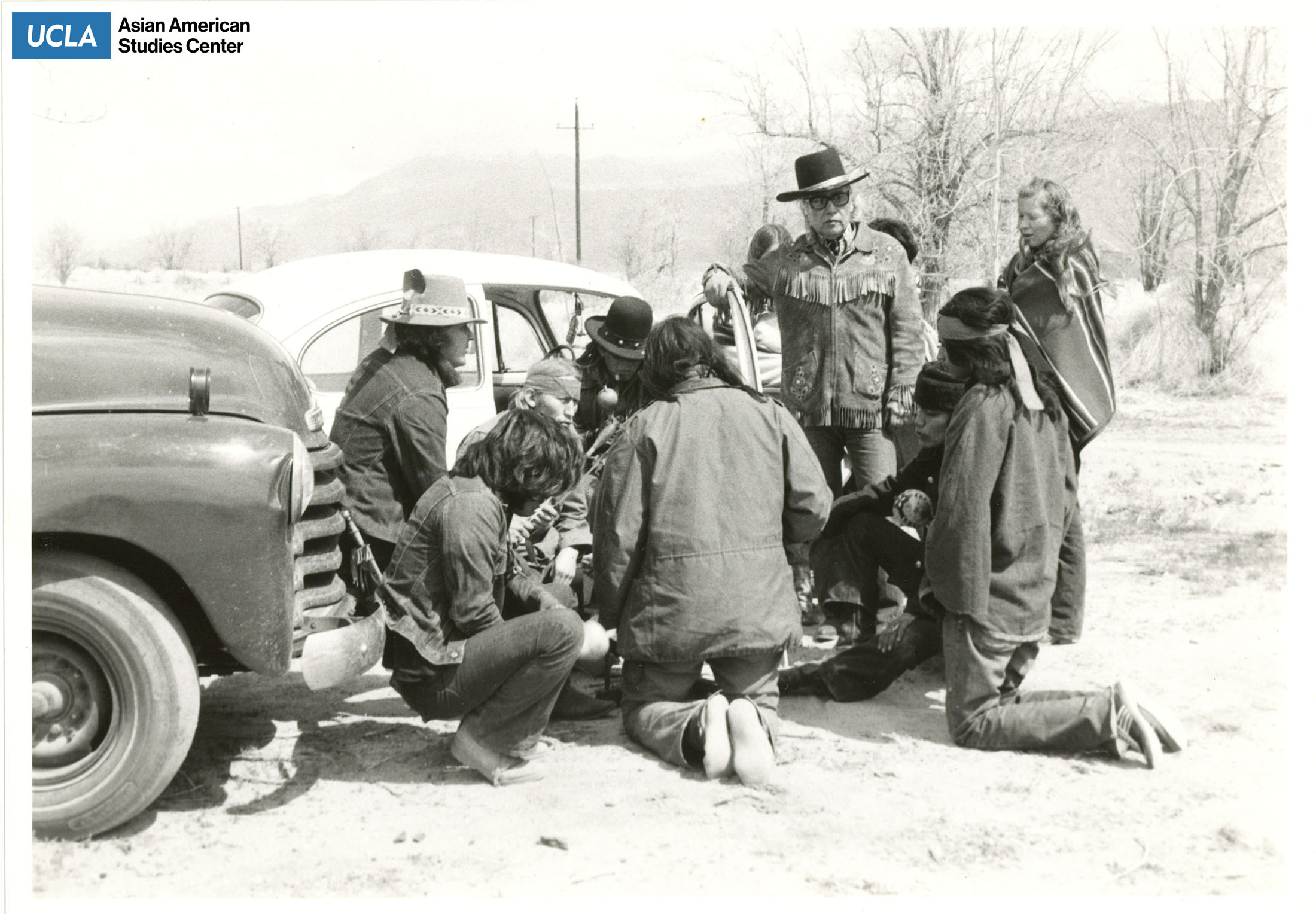 Black and white photograph of men sitting and standing in a circle with Volkswagen Beetle in background.